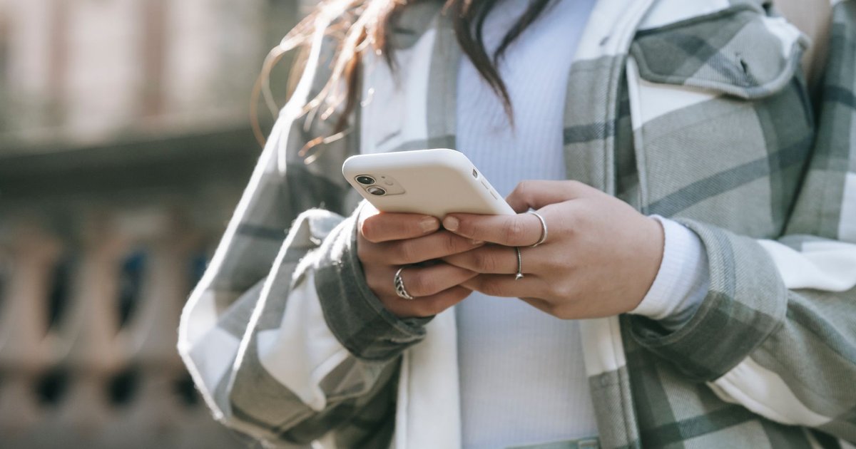 Jeune femme en chemise à carreaux tenant un smartphone blanc à deux mains dans une rue ensoleillée