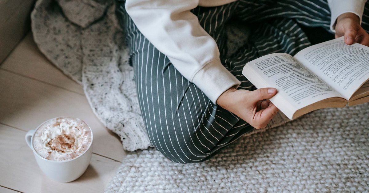 Lectrice assise en tailleur sur un tapis clair, un roman ouvert à la main et un chocolat chaud posé à côté