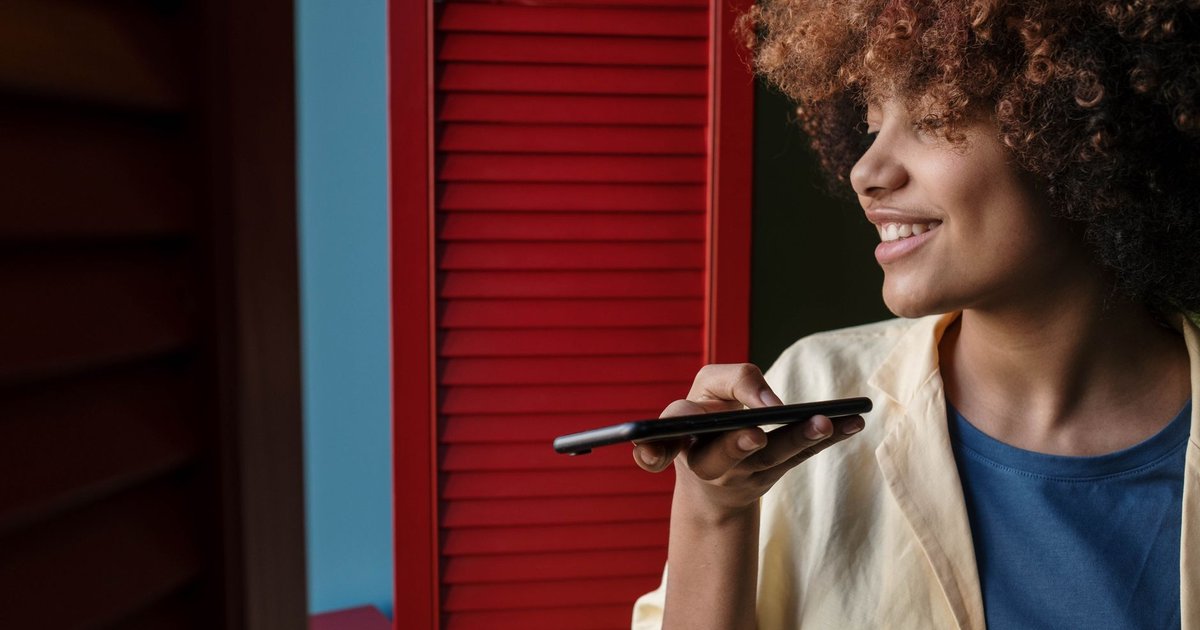 Jeune femme souriante qui dicte une note vocale à son téléphone, près d'une fenêtre aux volets rouges