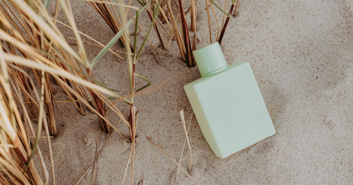 Flacon de parfum vert pâle posé dans le sable entre des herbes sèches