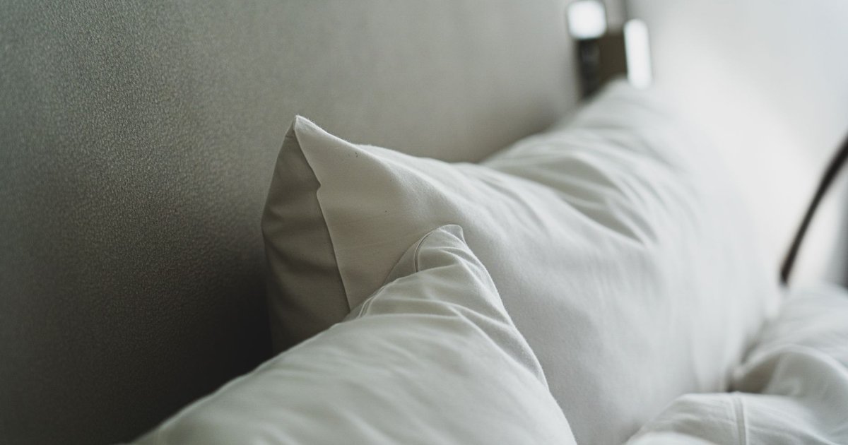 Oreillers blancs et couette froissée posés contre une tête de lit grise, dans une chambre baignée de lumière douce