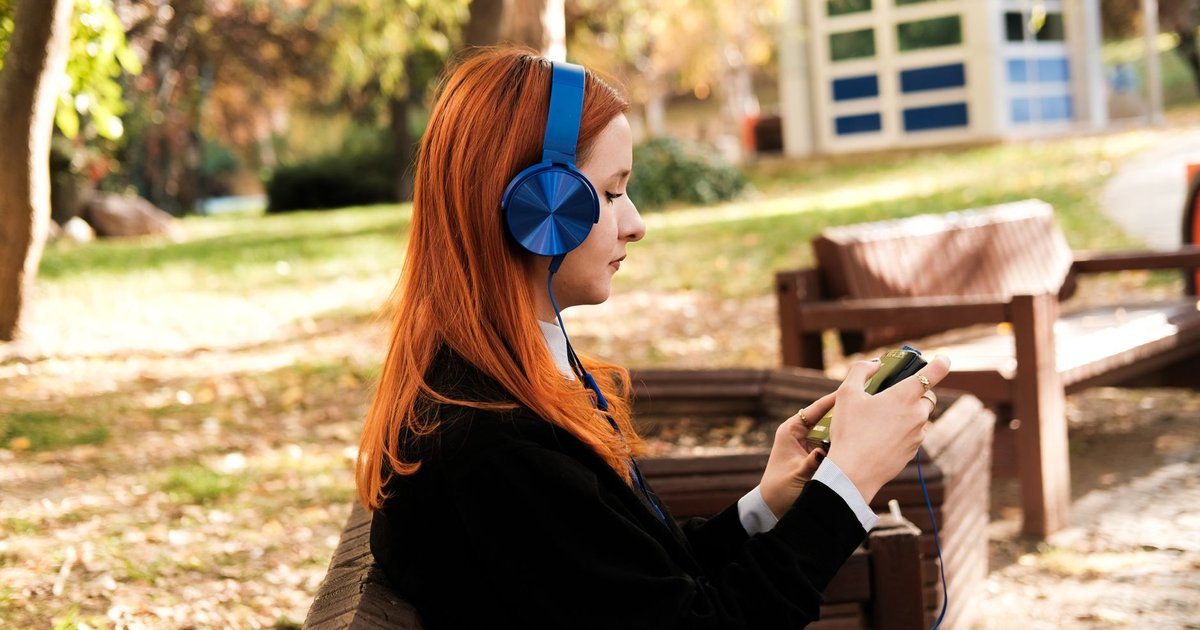 Jeune femme aux cheveux roux assise sur un banc de parc en automne, casque bleu sur les oreilles, qui choisit un morceau sur son téléphone