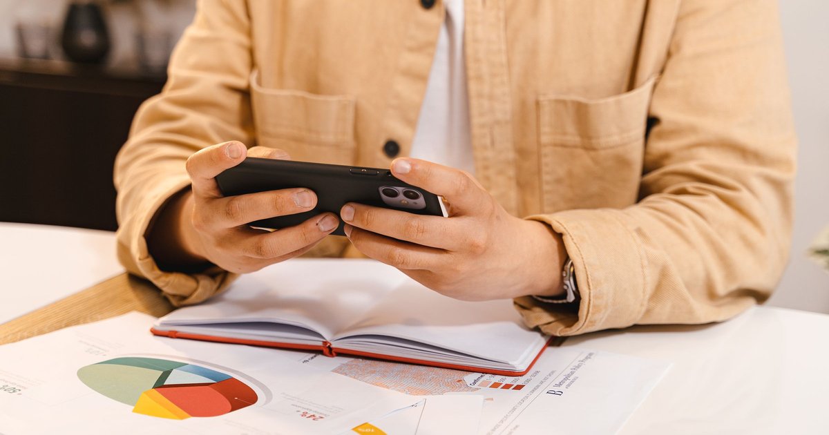 Mains tenant un smartphone au-dessus d'un carnet ouvert et de graphiques imprimés posés sur un bureau