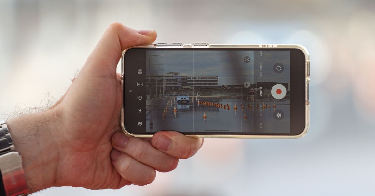Main tenant un smartphone qui filme un objet technologique posé sur un bureau clair