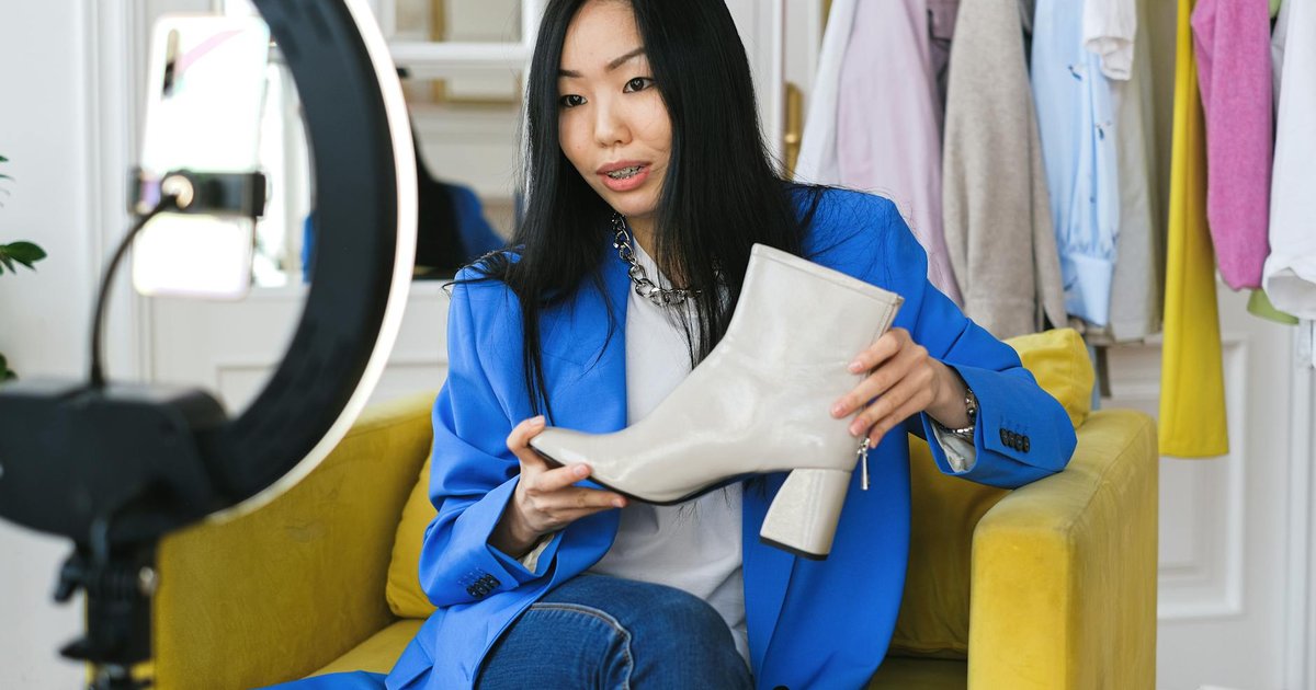 Créatrice en blazer bleu présentant une bottine blanche face à un smartphone monté sur un anneau lumineux, un portant à vêtements en arrière-plan lors d'un live shopping