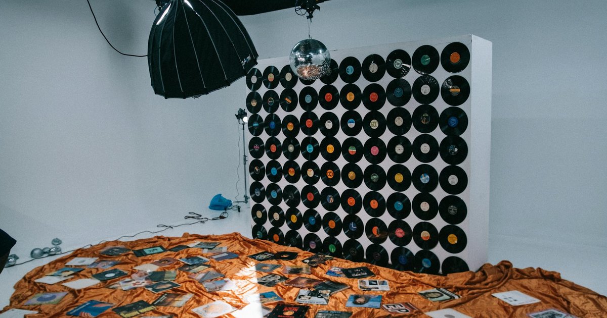 Mur de disques vinyles et boule à facettes dans un studio photo, pochettes d'albums éparpillées sur un tissu orange au sol